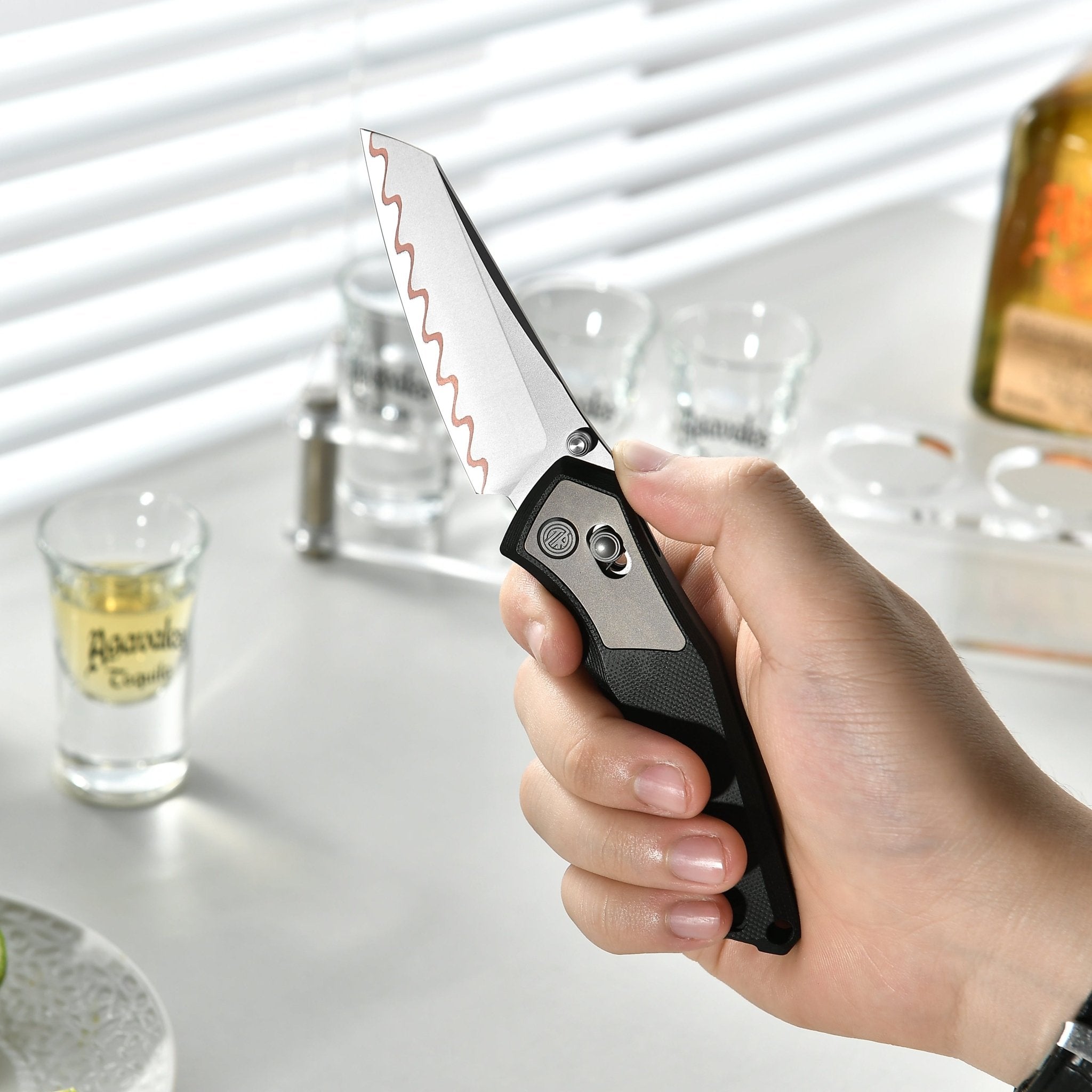 Hand holding Tacray Talos folding knife on a table with drinks and a cactus.